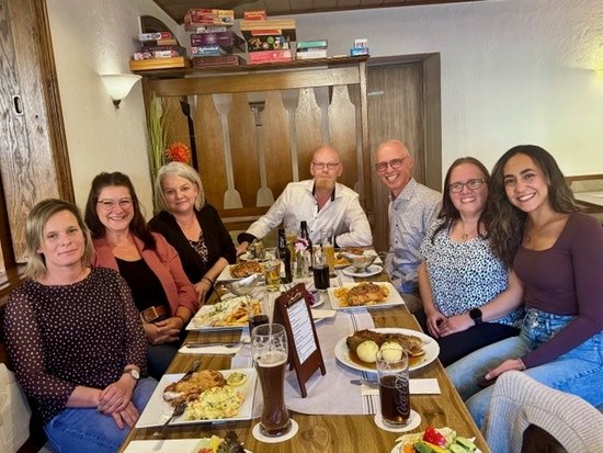 Sieben Personen sitzen lächelnd an einem langen Holztisch in einem Restaurant. Vor ihnen stehen Teller mit Essen, darunter Fisch und Pommes, sowie diverse Getränke. Im Hintergrund ist eine hölzerne Wanddekoration zu sehen.